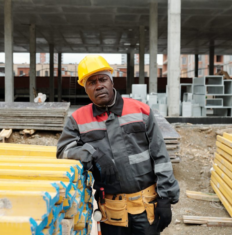 Serious mature black man in workwear and protective helmet looking at camera while standing by stack of yellow building materials